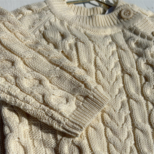 Close-up of a Essentials Cable Romper cream-colored cable knit sweater on a hanger.