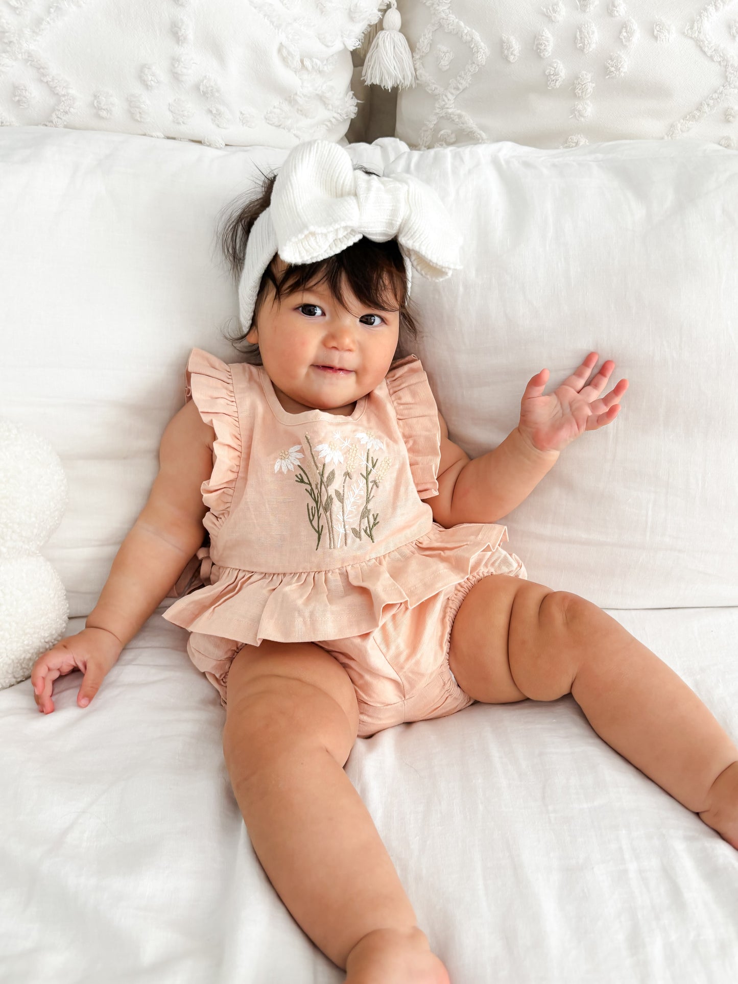 Baby in a pink romper with floral design and beige leggings lying on a white surface.