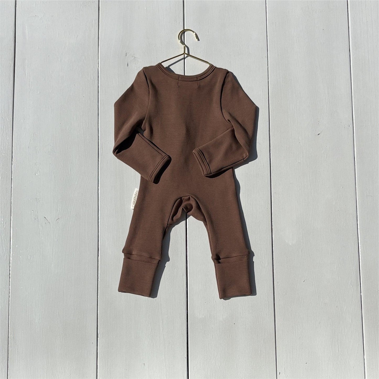 Brown organic baby onesie hanging on a gold hanger against white wood