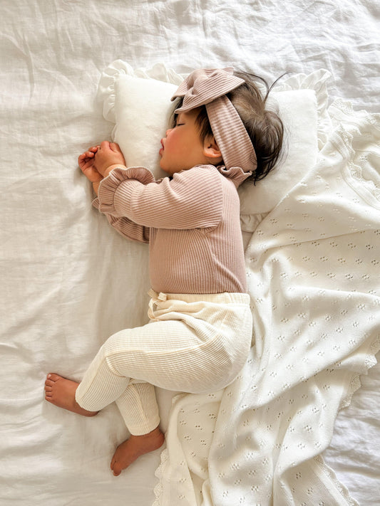 Baby wearing cream textured organic cotton pants with a beige long-sleeve top and headband on white bedding, Casita Bebes.
