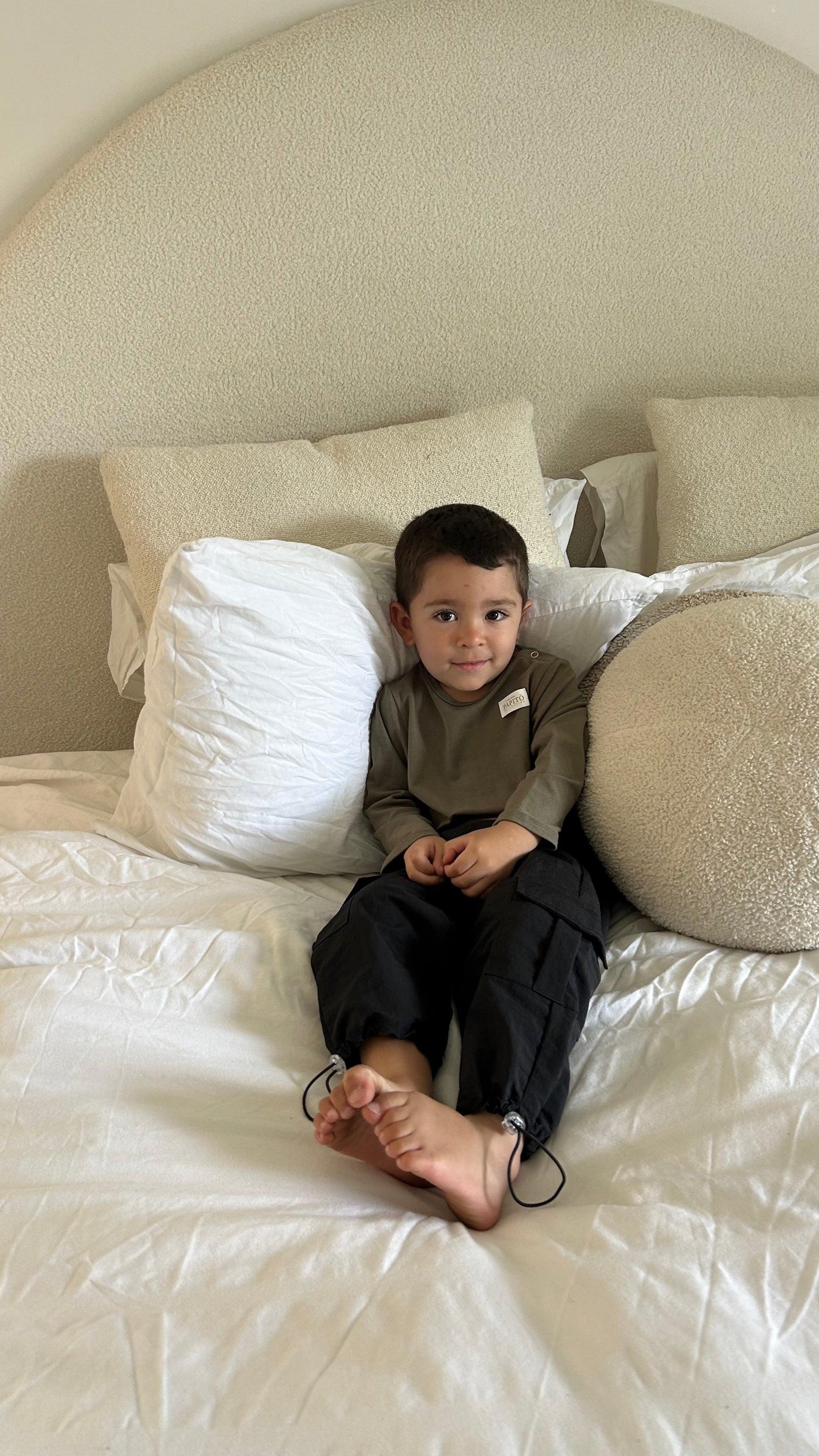 Toddler in khaki organic bamboo long-sleeve bodysuit sitting on cream bed, certified organic baby clothing.