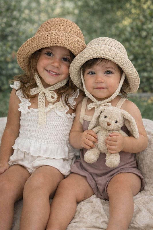 Two young children wearing bebe essentials crochet hats sitting outdoors with a blurred green background