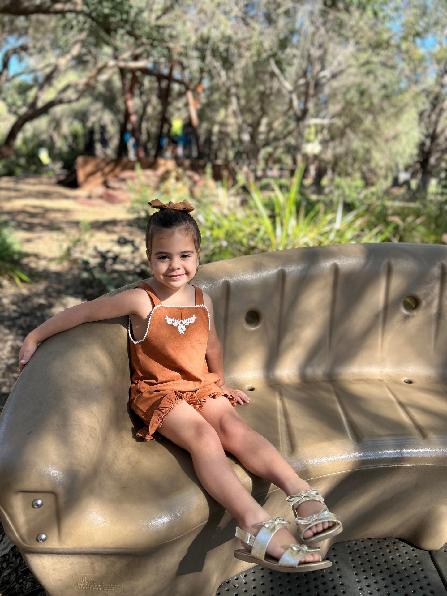 Toddler girl wearing certified organic Mamīta Sienna playsuit in rusty orange with bow detail, sitting outdoors.