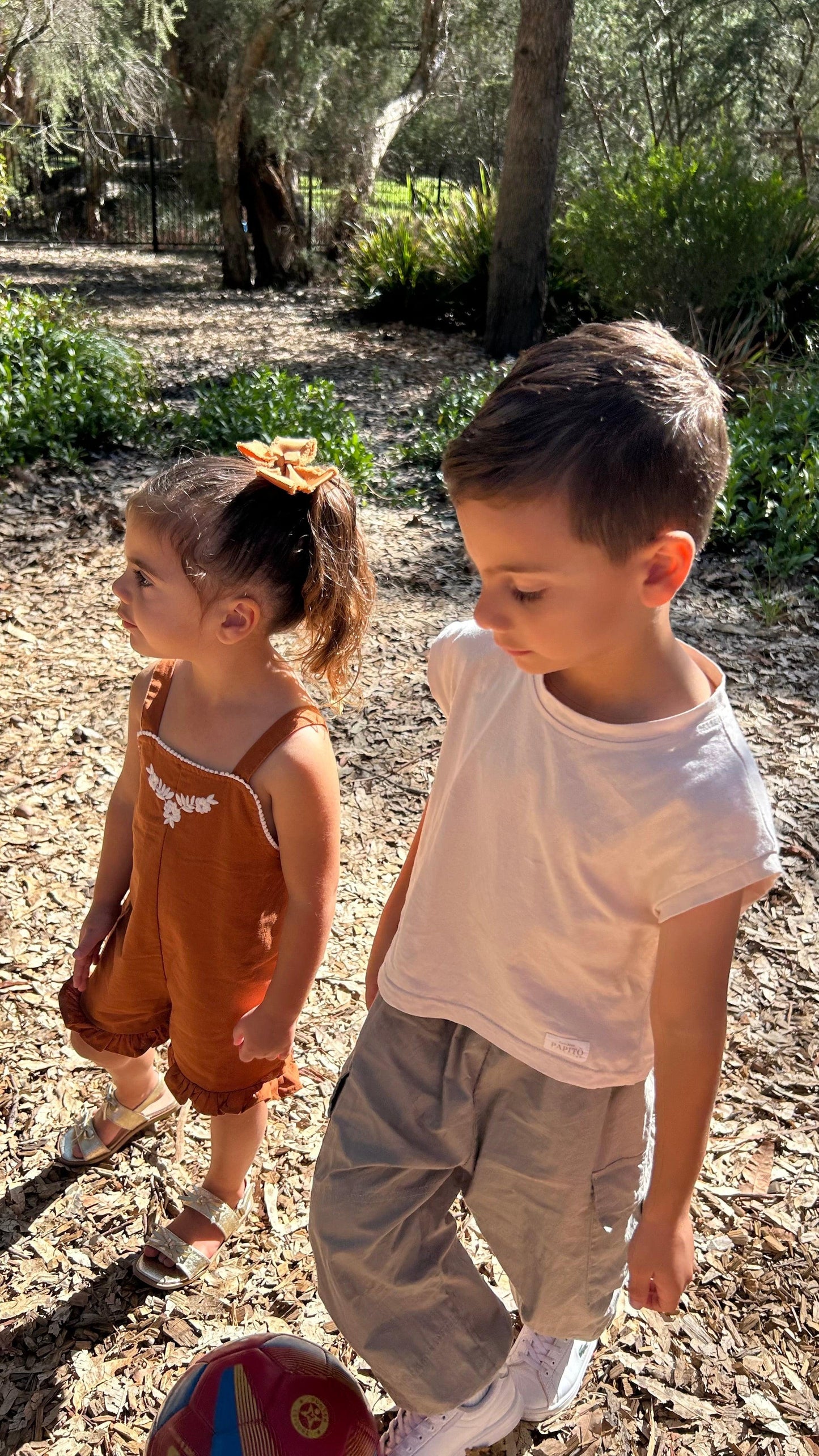 Toddler girl in organic rust Mamīta Sienna Playsuit with bow headband outdoors.