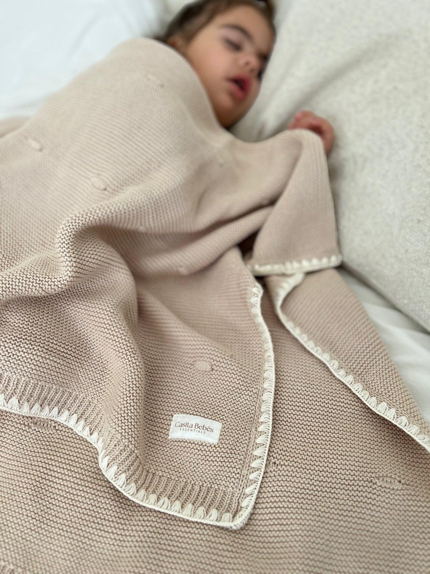 Child sleeping under a brown  knitted blanket with a visible label Casita Bebes