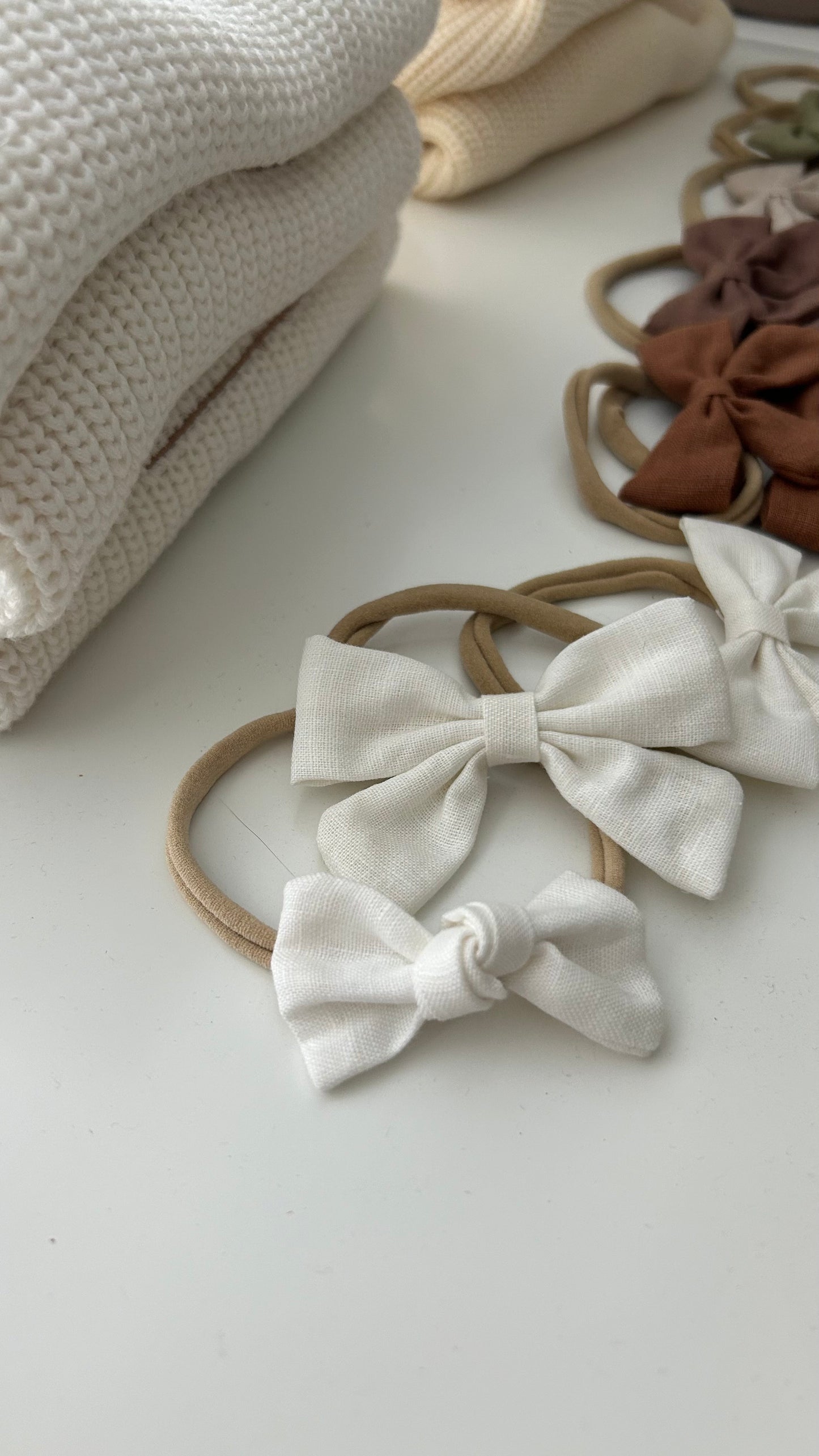 Two White fabric bows one small one big on brown hairbands with beige fabric in the background casita Bebès brand