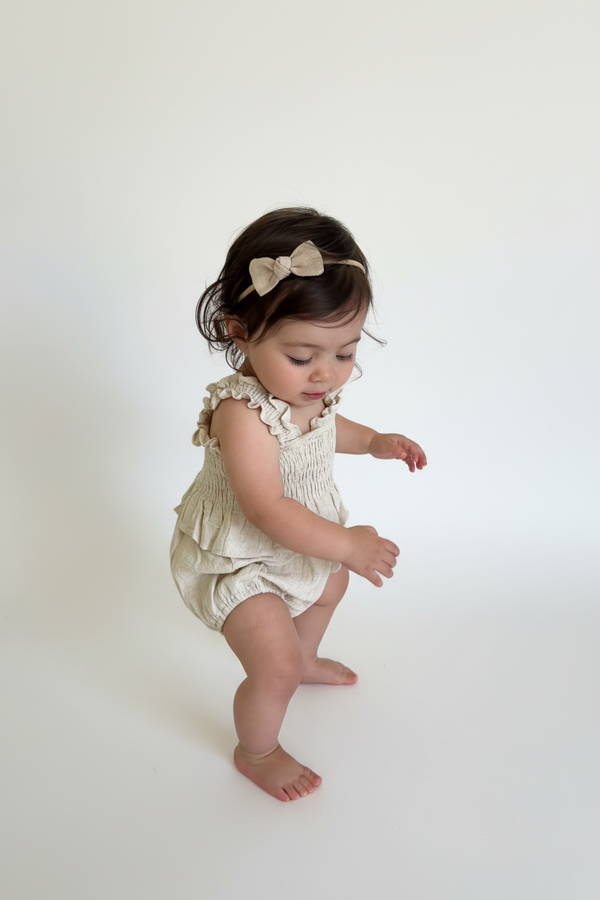 Baby with linen bow in a beige rcasita bebes romper with ruffles standing on a white background