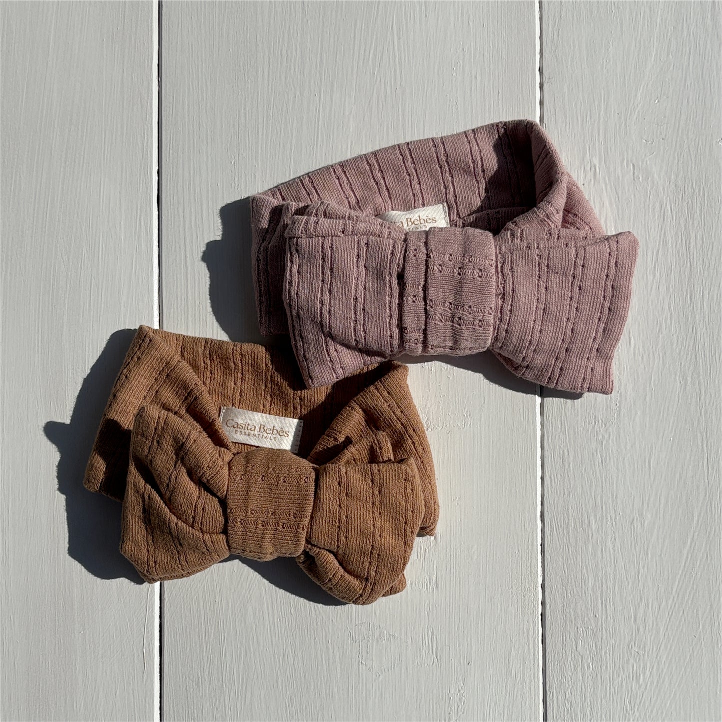 Two knitted headbands, one brown and one dusty purple, on a light wooden surface.