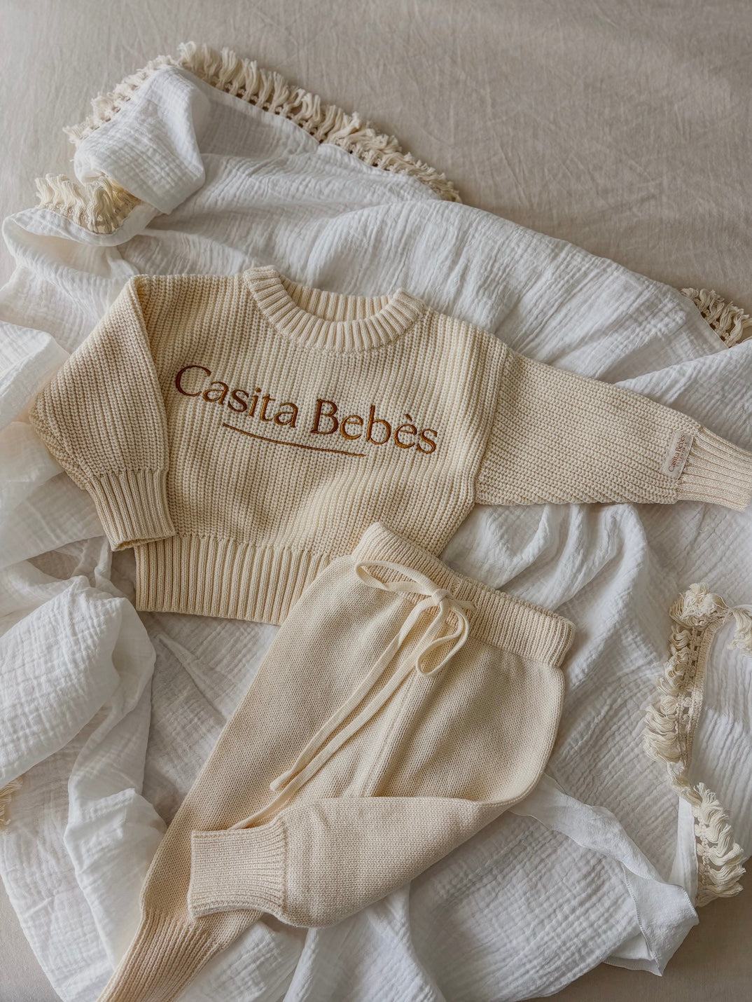 Beige baby sweater and pants flat lay with 'Casita Bebes' text on a textured surface.