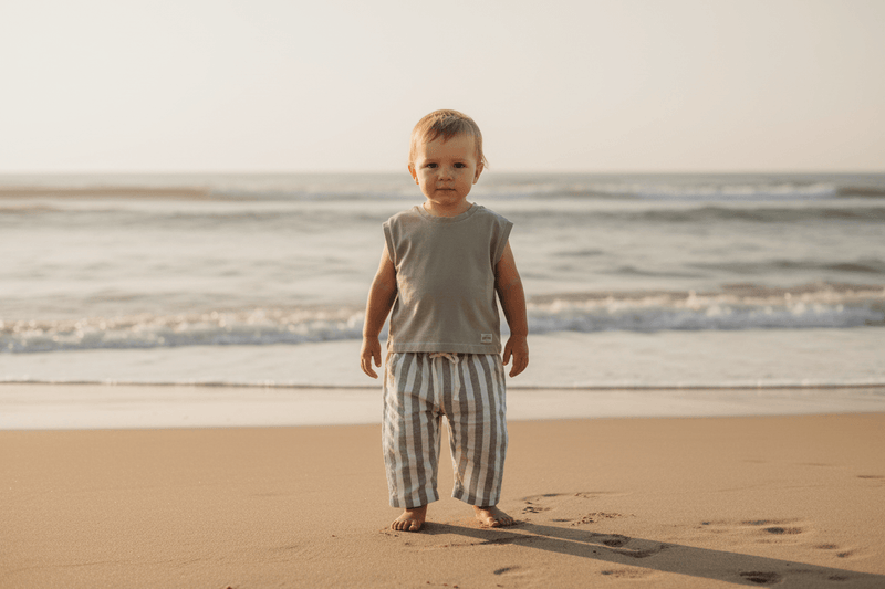 Baby in organic striped pants and sleeveless top standing on beach at sunset