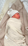 Newborn baby swaddled in a beige blanket with a light gray hat, lying on a textured beige blanket.