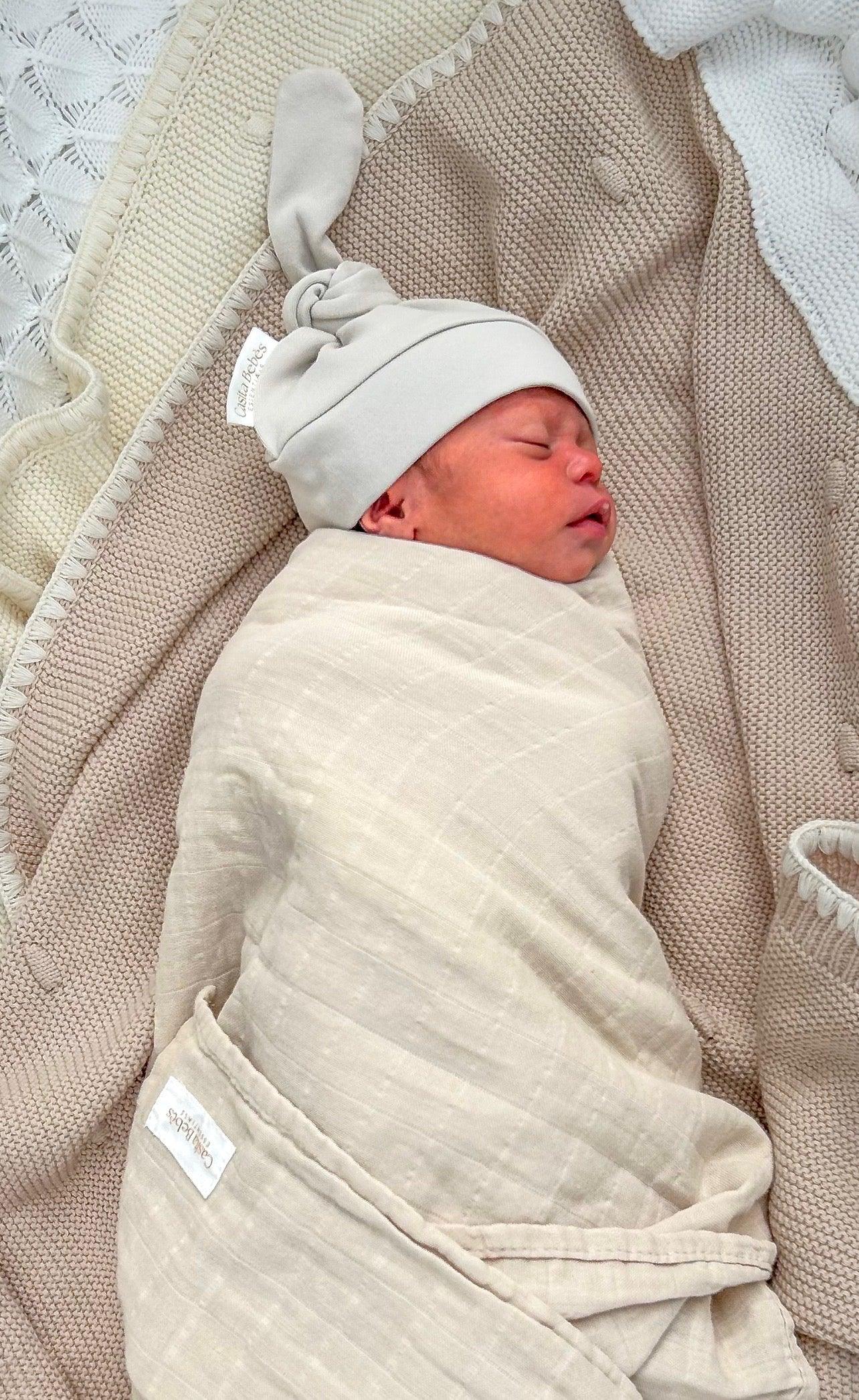 Newborn baby swaddled in a beige blanket with a light gray hat, lying on a textured beige blanket.