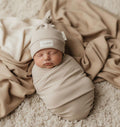 Newborn baby swaddled in organic cotton wrap with matching beanie, nestled on soft blanket