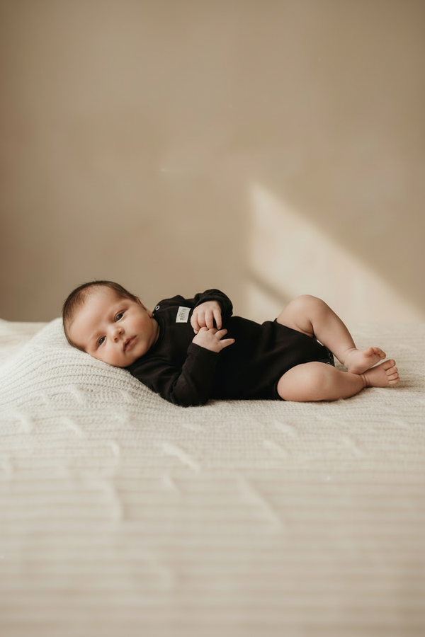 Newborn baby in a black onesie lying on a beige blanket with a soft light source. Casita Bebes