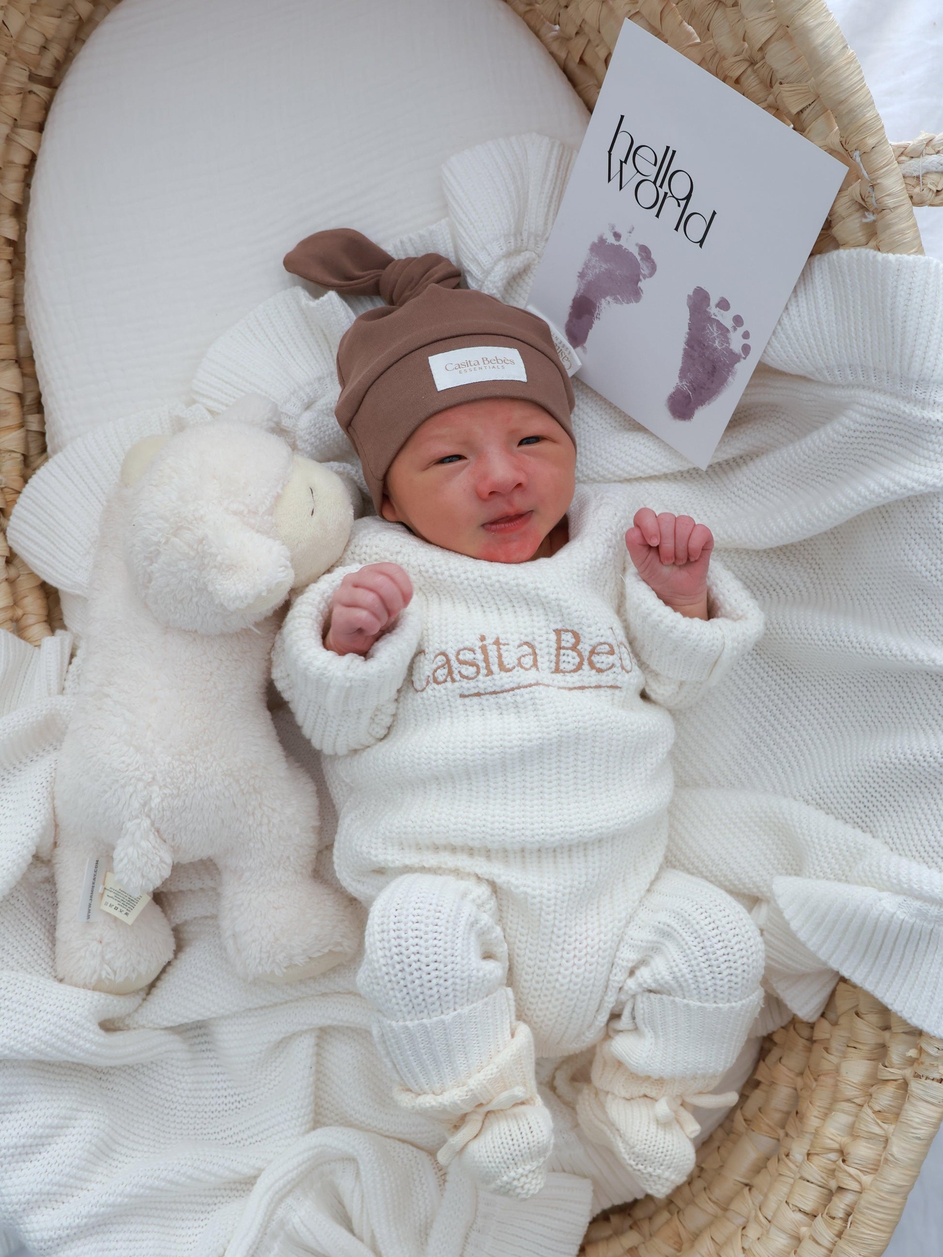 Newborn baby in a white outfit with 'Casita Bebes' branding, wearing a brown hat, surrounded by soft textures and a 'hello world' card.