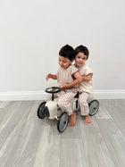 Two children playing on a small bike indoors with a plain white wall background casita bebes