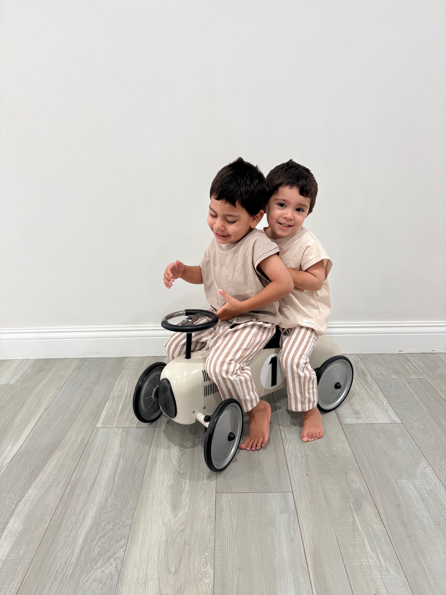 Two children playing on a small bike indoors with a plain white wall background casita bebes