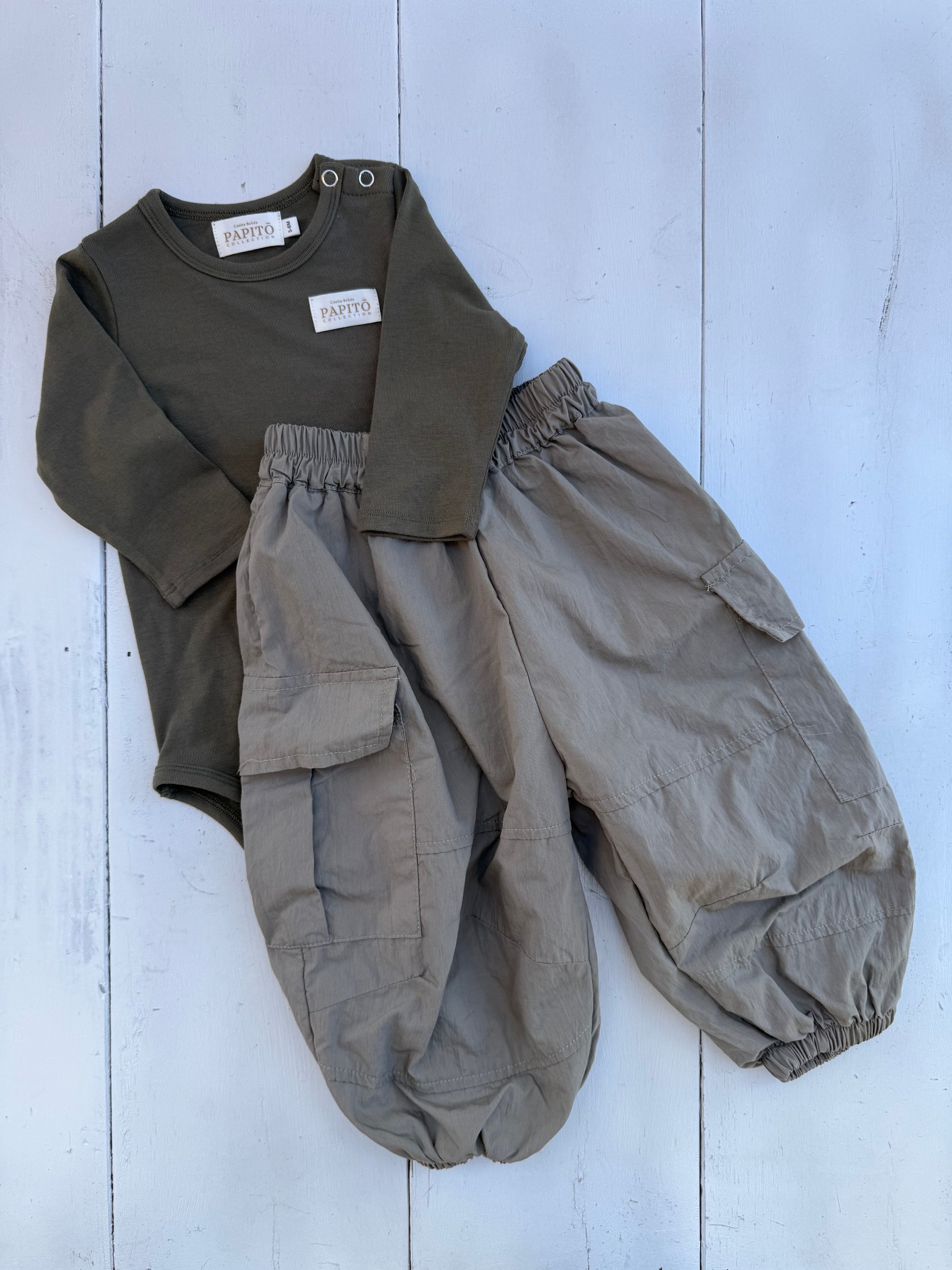 Children's outfit with green long-sleeve shirt and beige cargo pants on a light wooden floor. casita Bebes