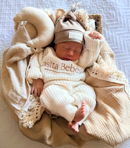 Newborn baby in a beige outfit with Casita Bebes' branding, wrapped in a blanket on a textured surface.