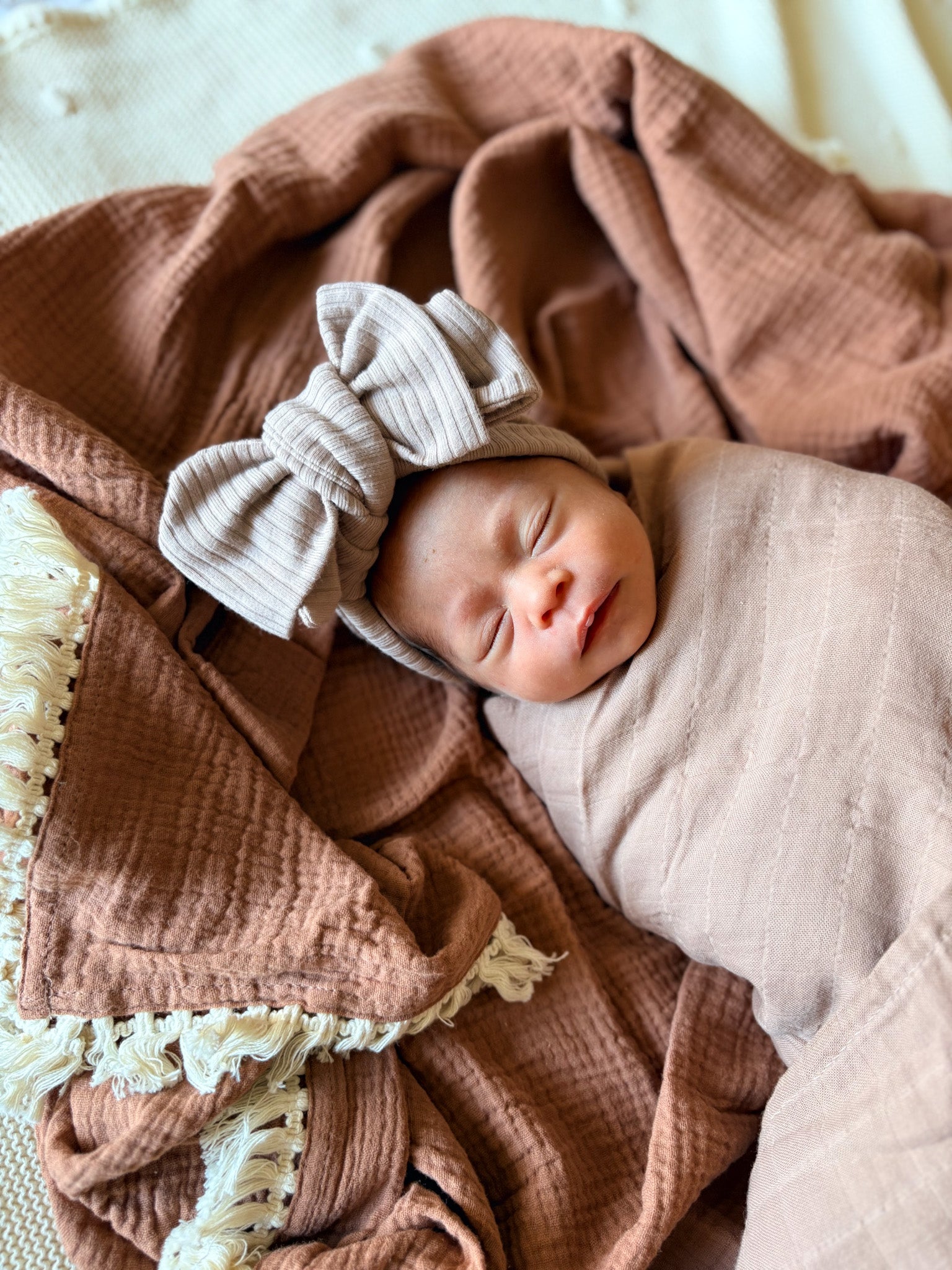 Newborn baby wrapped in a pink blanket with a matching bow headband, lying on a soft brown blanket.