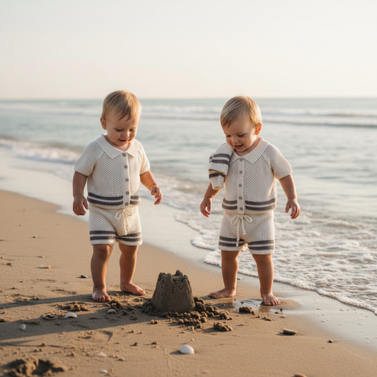 Two children in matching outfits standing on a beach with a sandcastle.