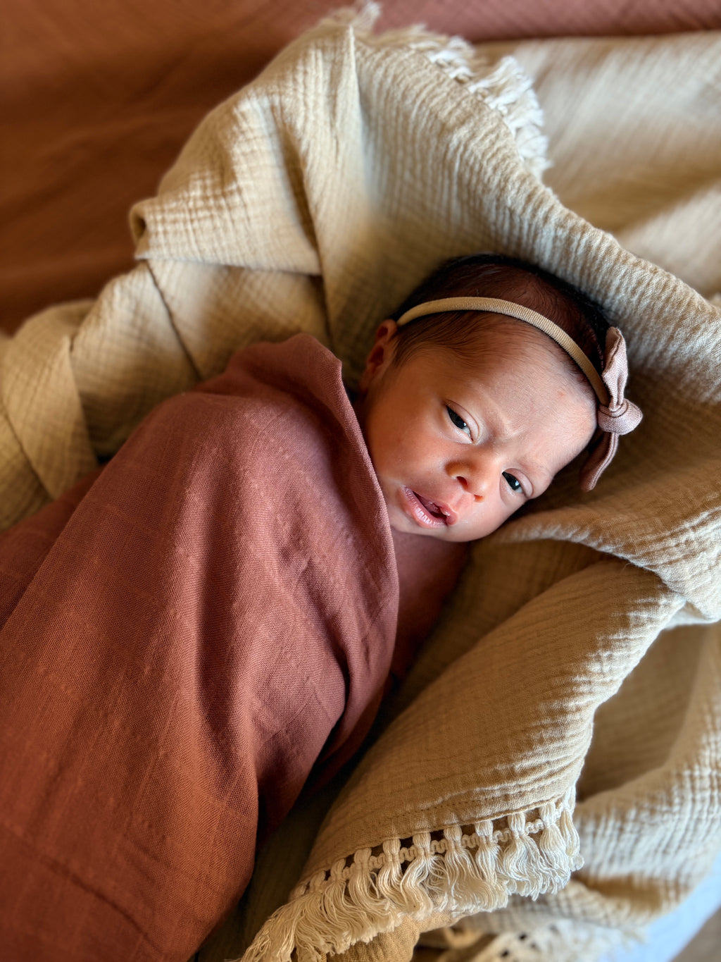 Newborn baby wrapped in a tuscan Mediterranean brown blanket with a beige headband, lying on a textured beige blanket. Casita Bebes