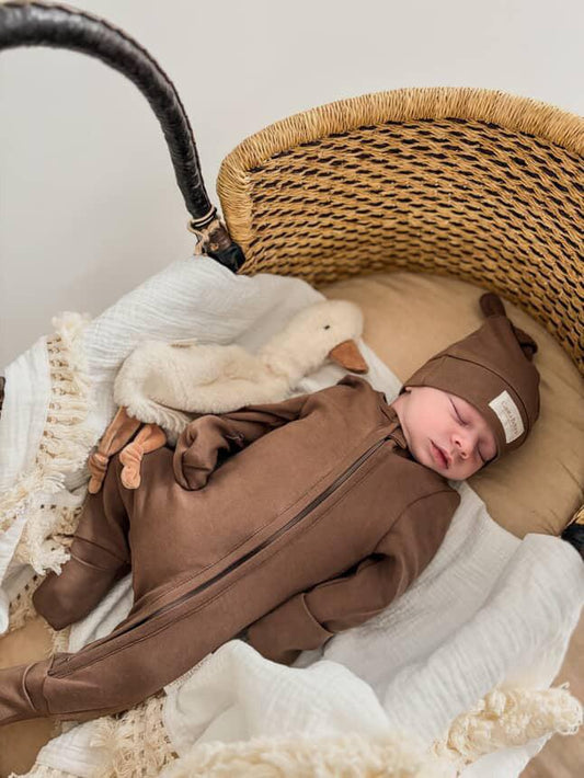 Sleeping baby wearing dusty brown organic romper and matching hat in woven bassinet with white textured blanket.