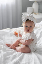 Baby in white ruffle onesie with bow headband, sitting on organic cotton blanket