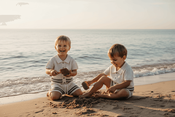 Two toddlers in organic knit outfits playing with sand by the ocean at sunset
