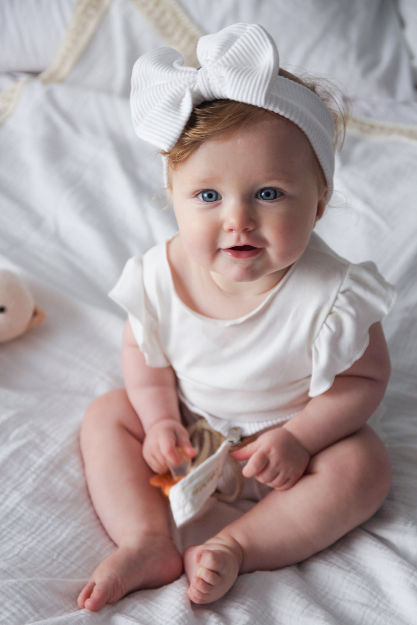 Baby wearing a white outfit and headband sitting on a white blanket.