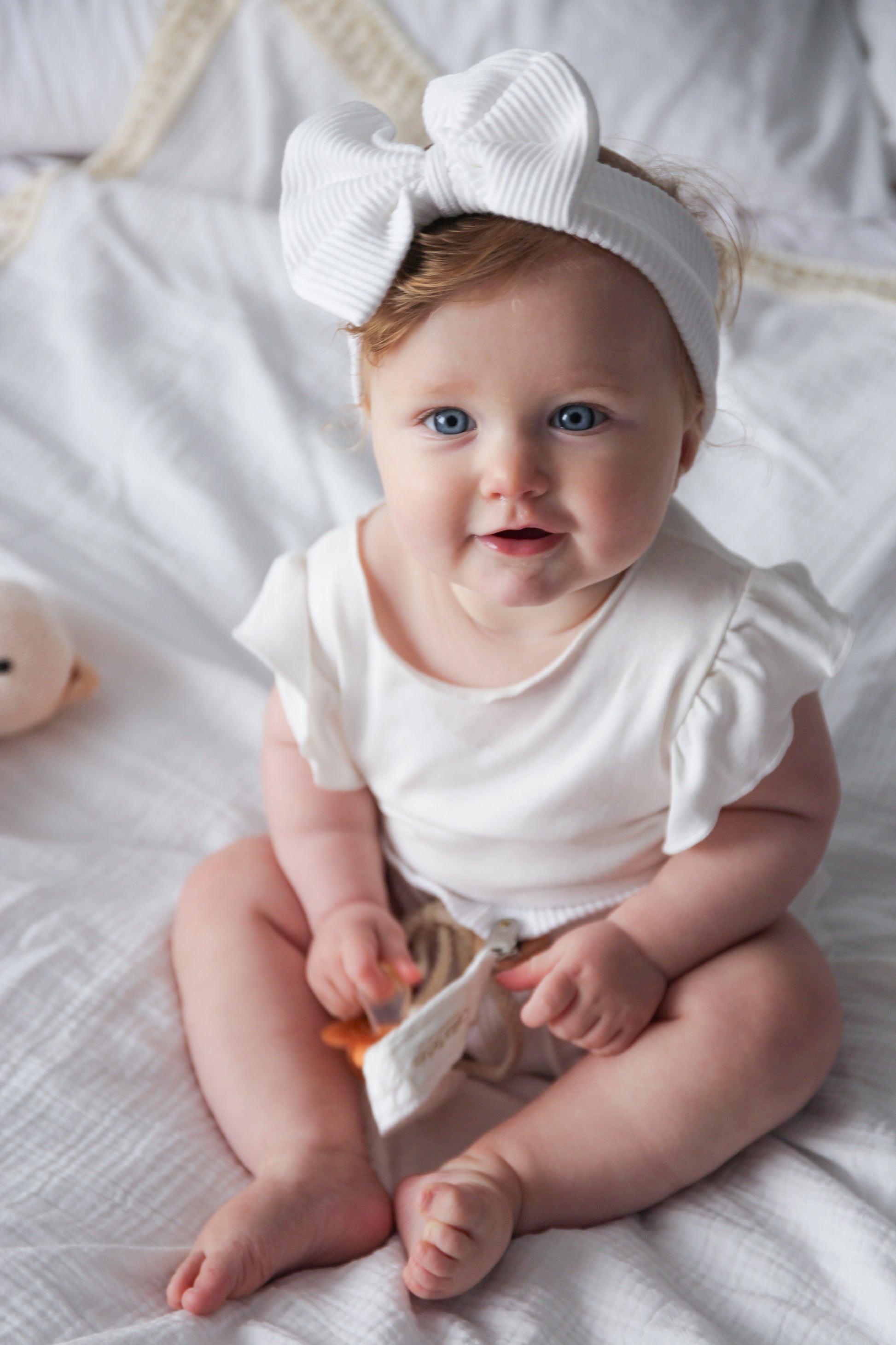 Baby wearing a white outfit and headband sitting on a white blanket.