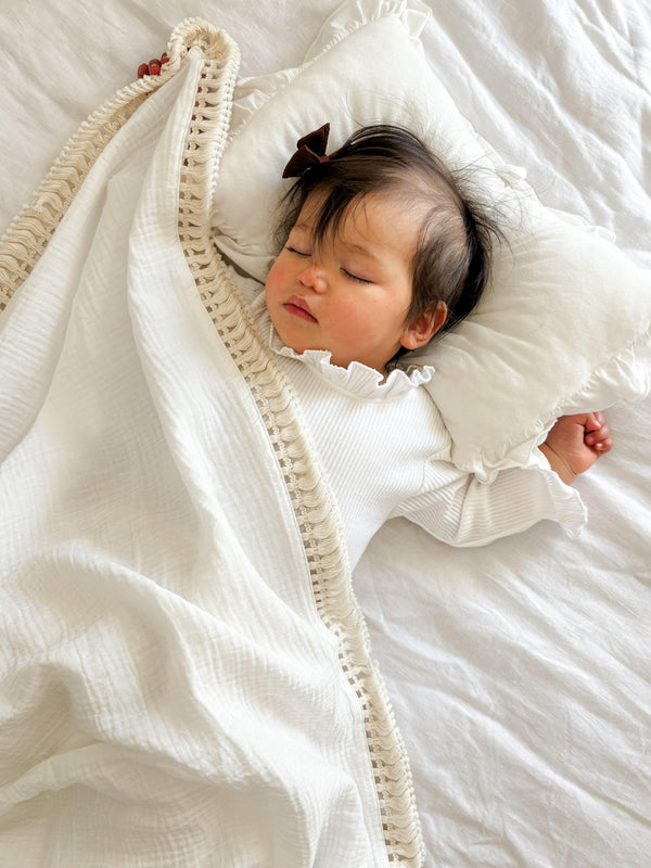 Certified organic cream cotton tassel blanket gently draped over sleeping baby wearing white clothing.
