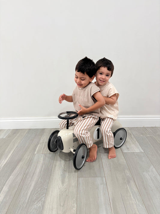 Two toddlers wearing dusty beige organic baby clothing riding a cream vintage toy car indoors.