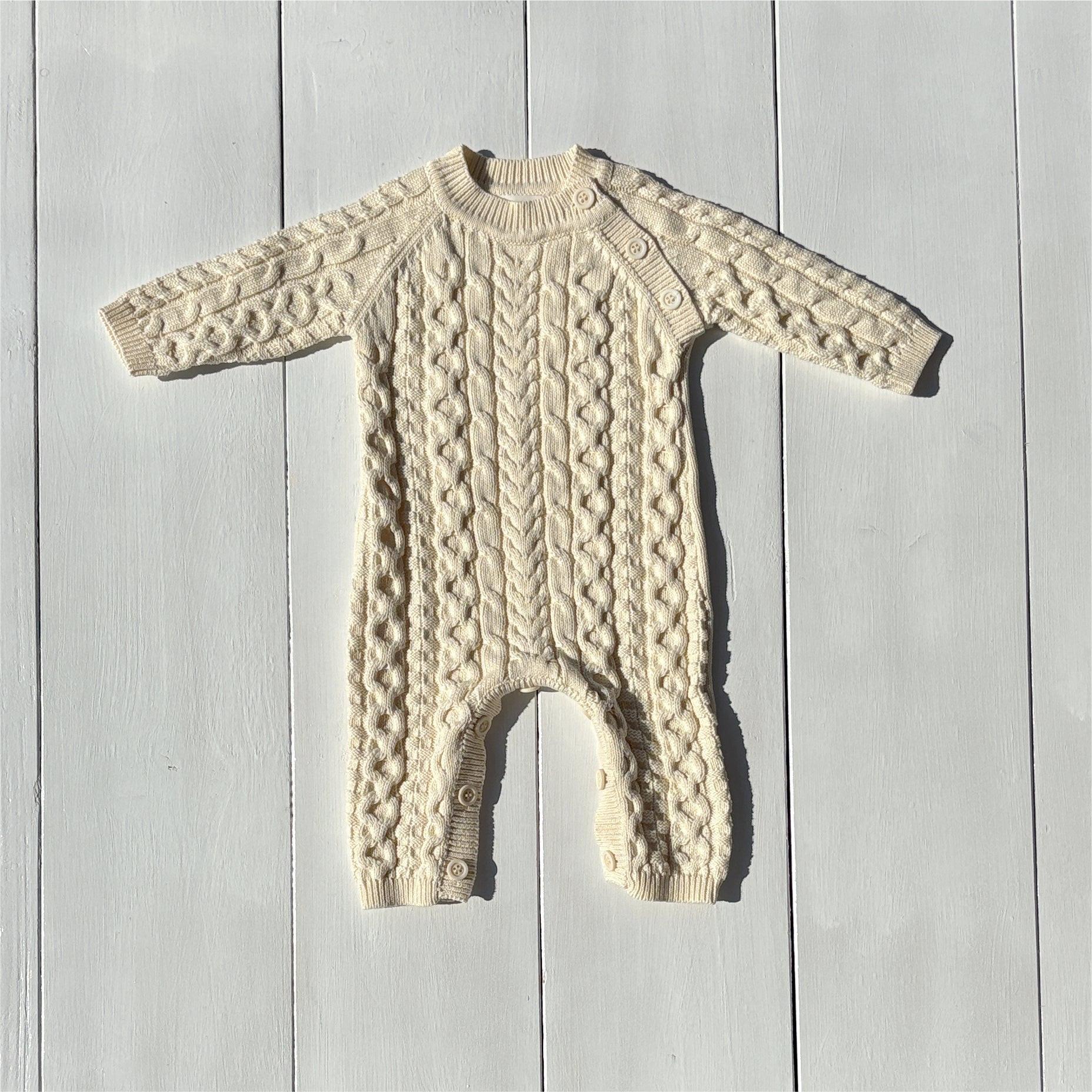 Cream-colored knitted organic baby romper with cable pattern, long sleeves, and certified organic fabric.