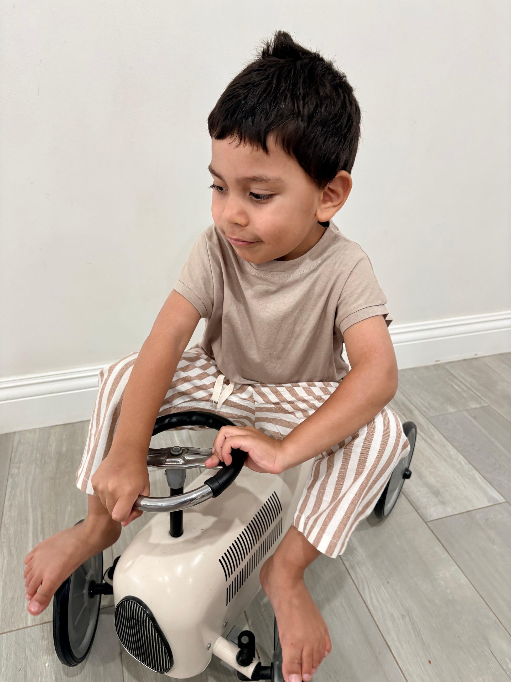 Toddler boy wearing dusty brown Papitō Milano organic baby pants with beige top, seated on toy car.