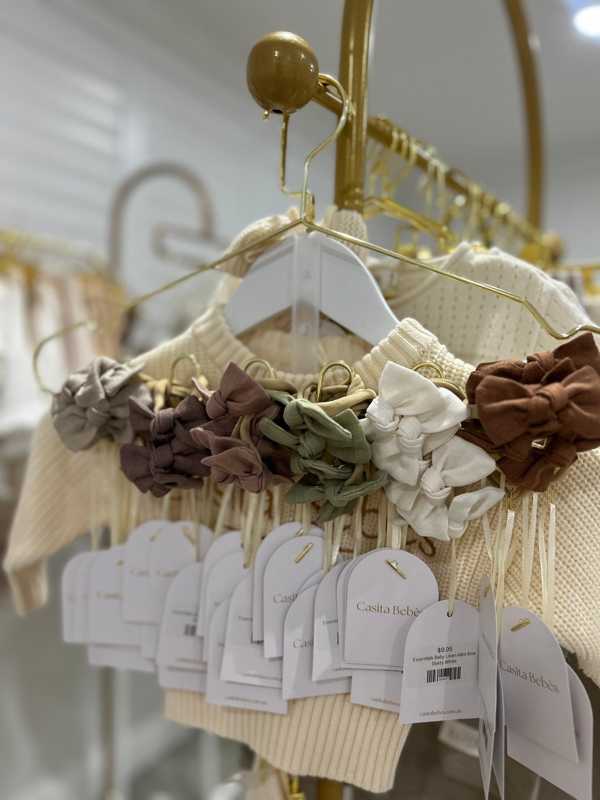Organic baby clothing with mini bows in neutral tones, displayed on gold hangers for gentle early motherhood essentials.