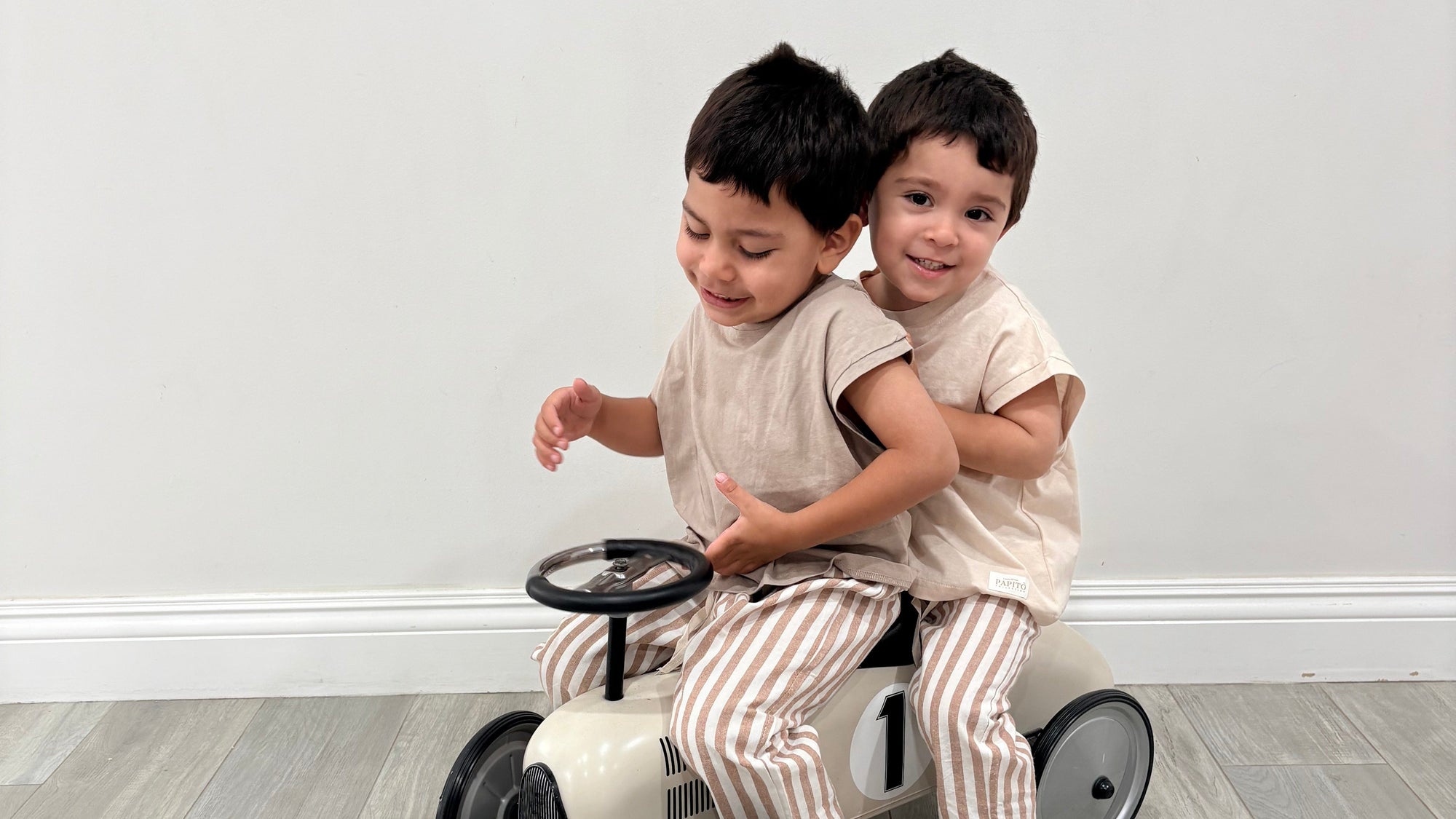Two children playing on a small bike indoors with a plain white wall background