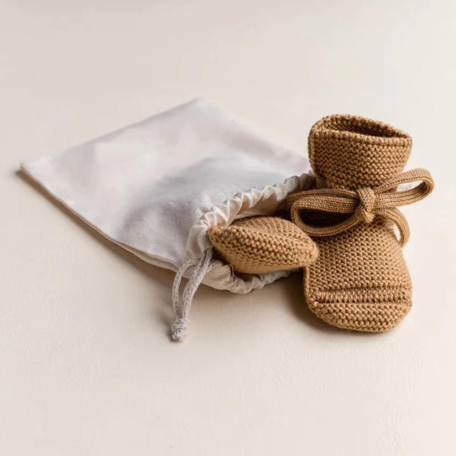 Organic dusty brown knit baby booties with ties beside white drawstring pouch, certified organic baby clothing by Casita.