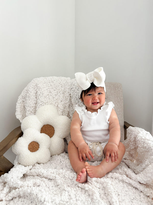 Baby in white ruffled top and bow headband sitting on cozy blanket with daisy pillow, organic baby essentials