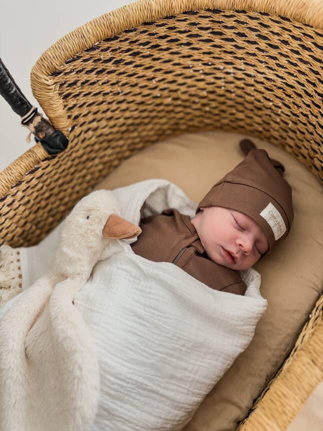 Sleeping baby wearing dusty brown organic knot beanie and matching baby clothing in woven bassinet.