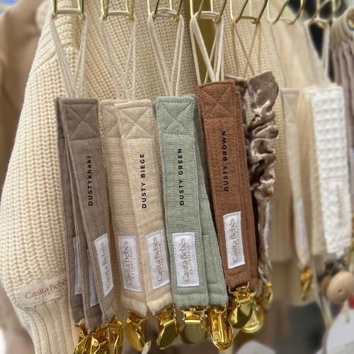 Set of certified organic Essentīals Gold Dummy Holders in dusty beige, brown, green, and khaki hanging on baby clothing rack.