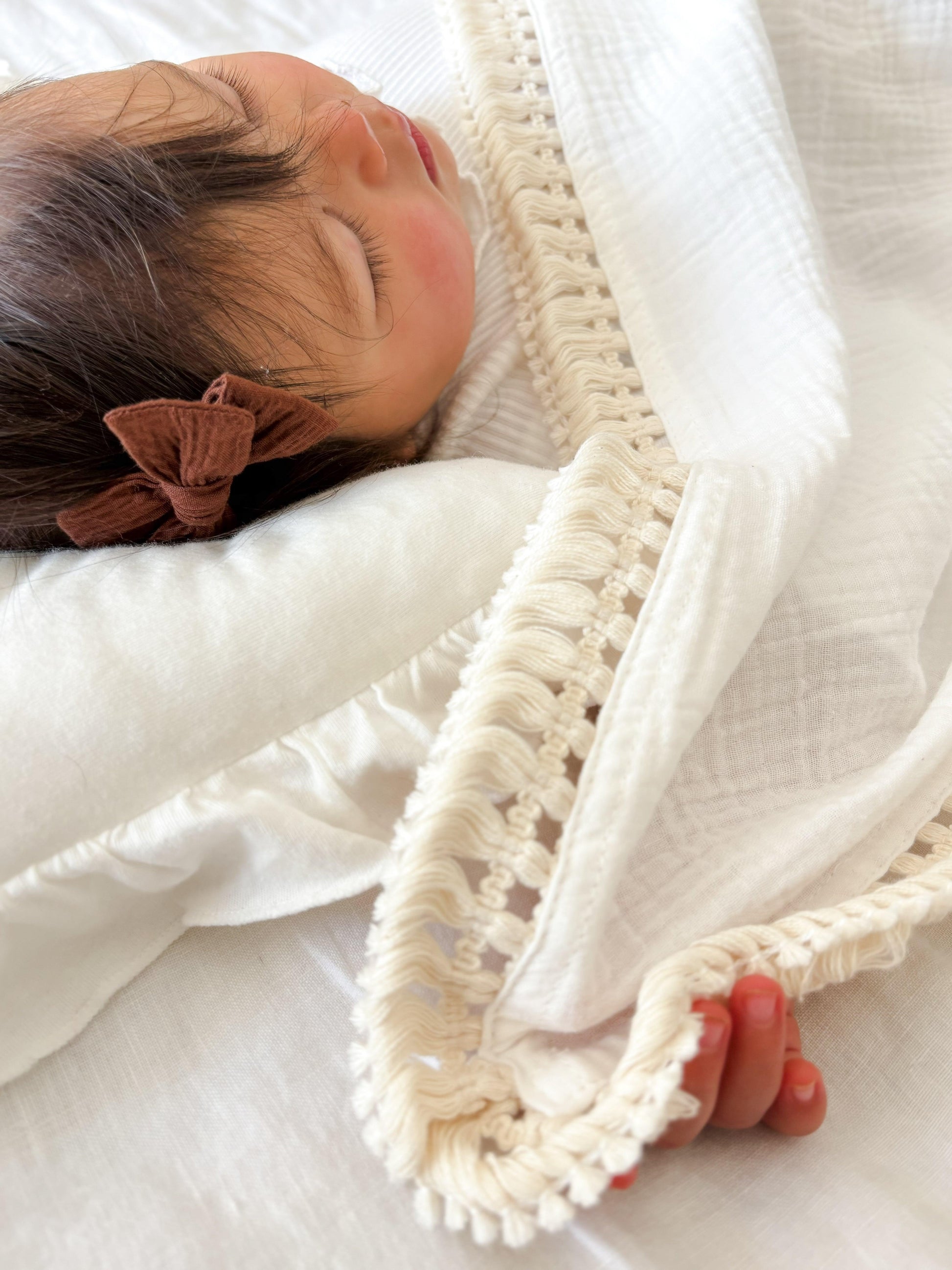 Baby sleeping peacefully under organic cotton blanket with macrame trim, wearing brown bow headband