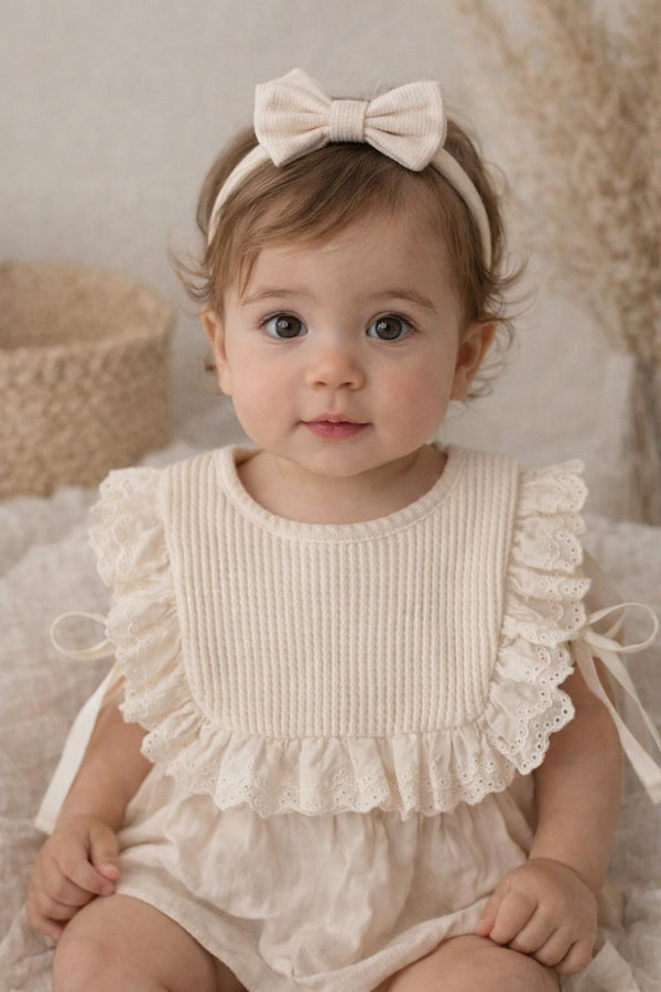 Baby toddler wearing a white ruffled bib and headband with a bow, sitting on a soft surface.