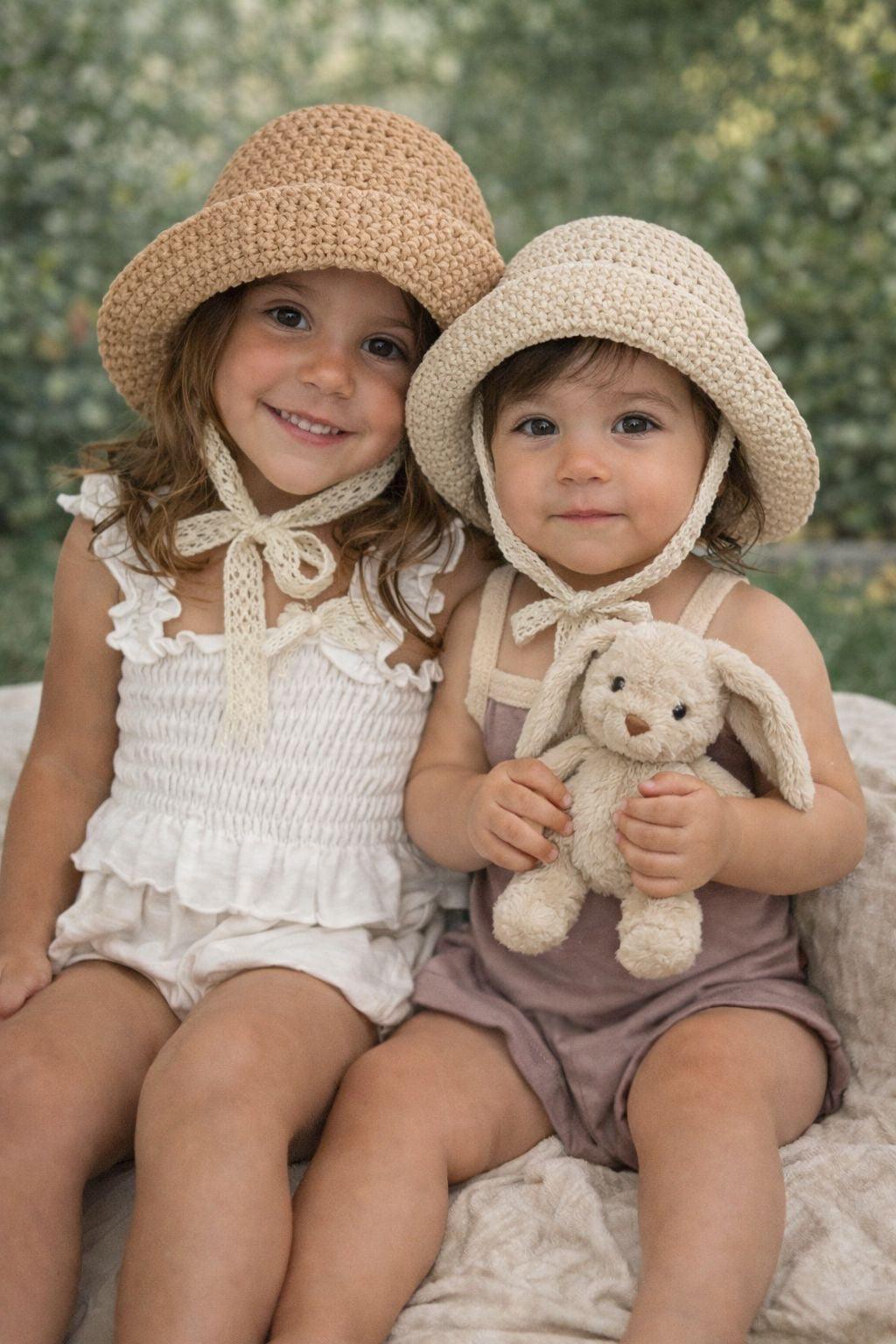 Two children in organic baby clothing, wearing crocheted sun hats, one holding a plush bunny toy.